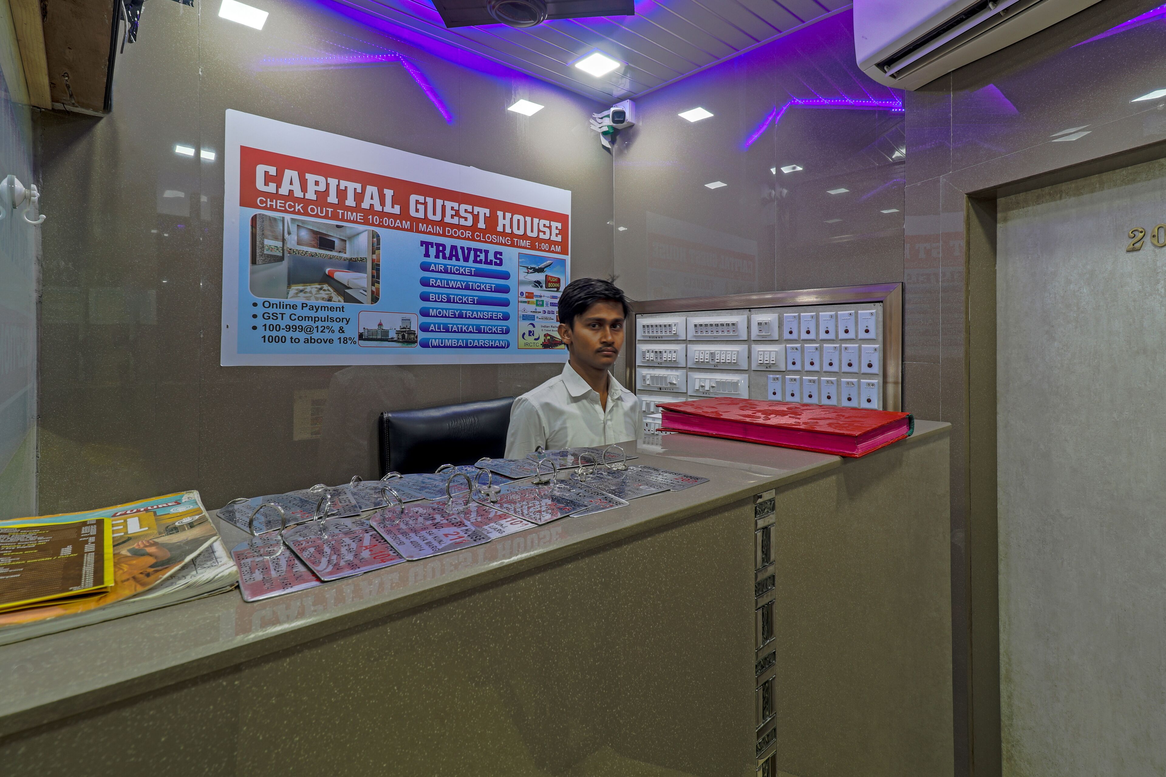A reception desk with a signboard for Capital Guest House, a man sitting behind it, and a wall-mounted air conditioning unit.