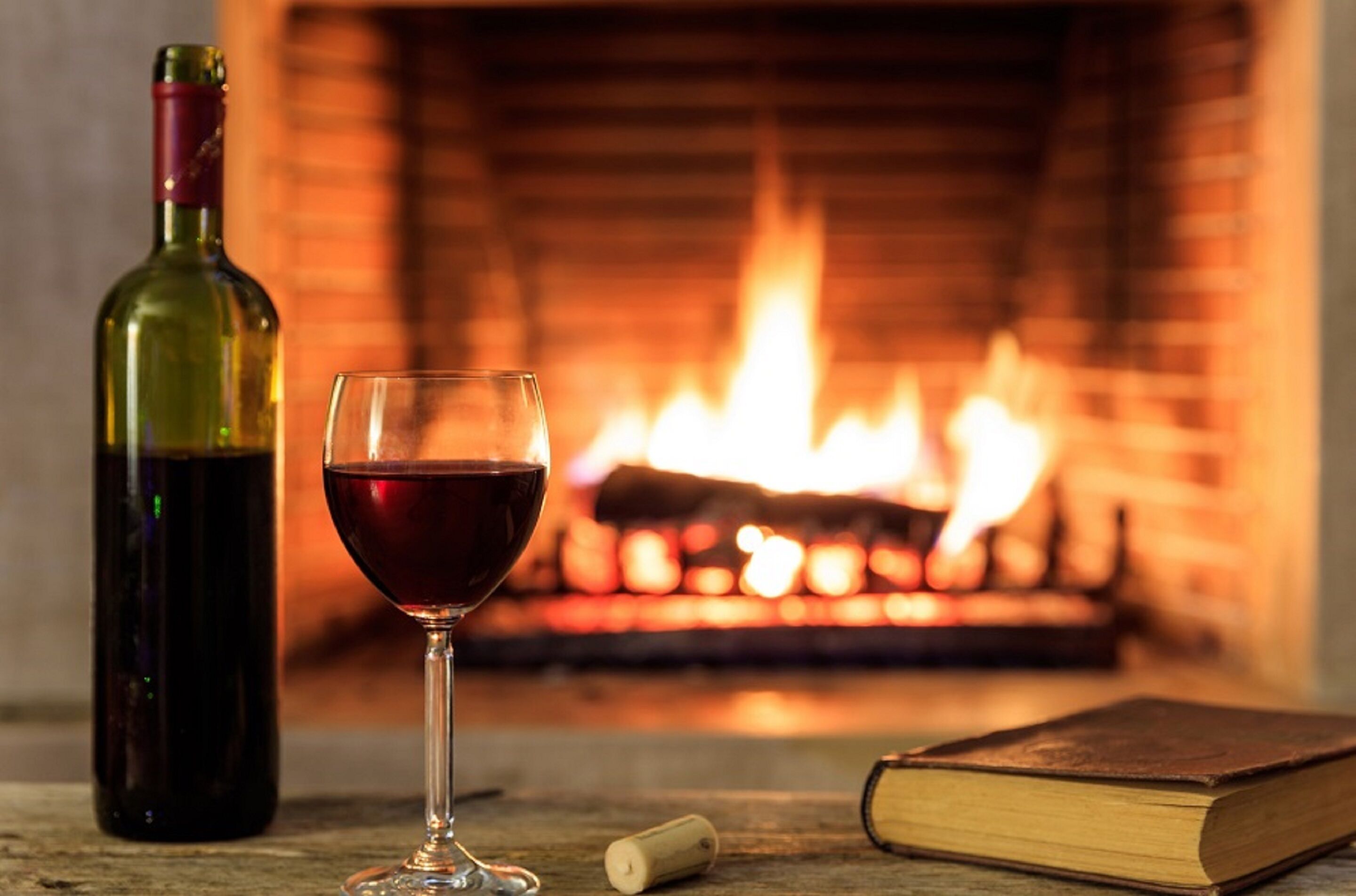 A bottle of wine, a glass of red wine, a cork, and a book in front of a fireplace.