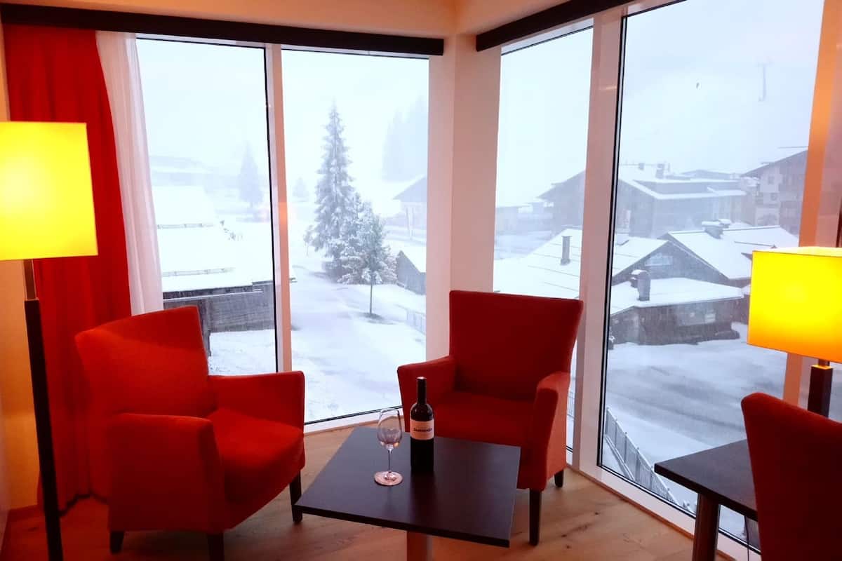 Une chambre d’hôtel avec deux fauteuils rouges, une petite table sur laquelle se trouvent une bouteille de vin et un verre, et de grandes fenêtres donnant sur un paysage enneigé.