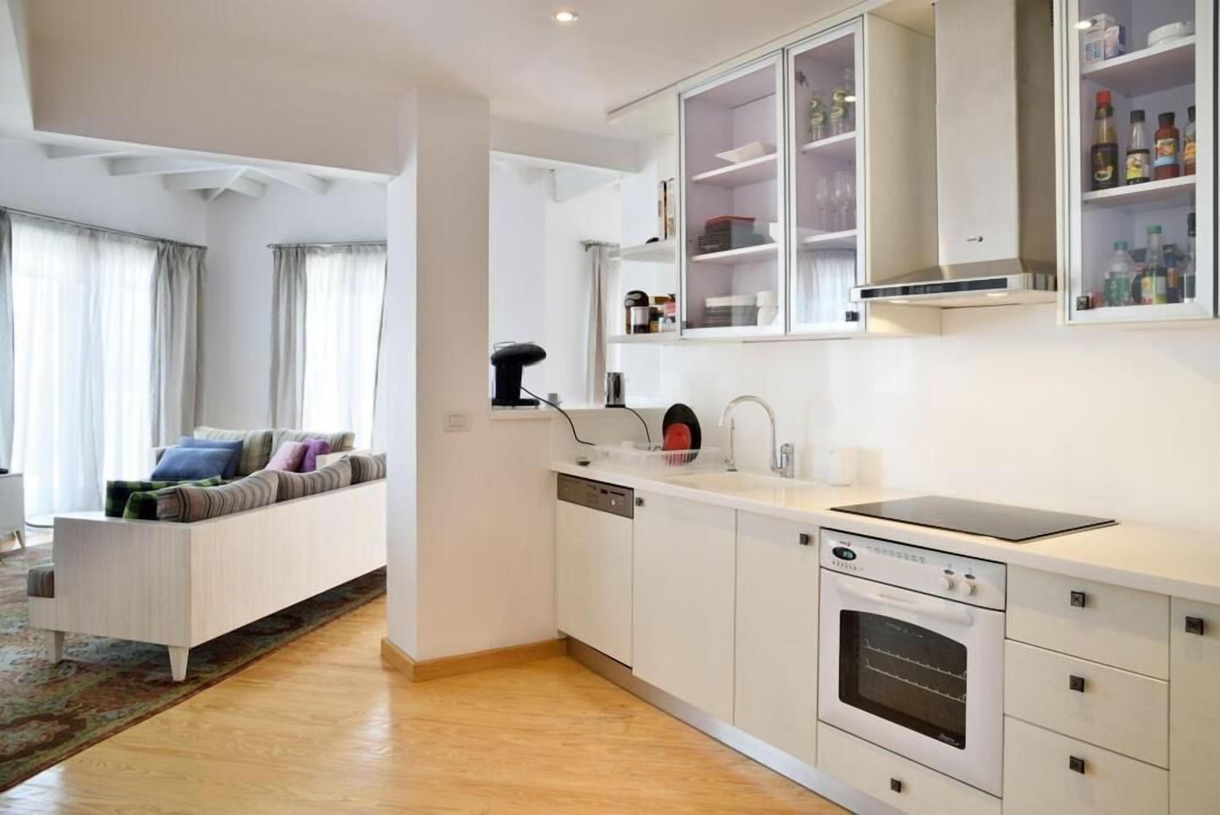 Una cocina moderna con armarios blancos, horno integrado y un comedor con sofá y cortinas.