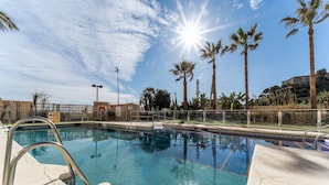 Una piscina con una escalera metálica, rodeada de palmeras y un cielo despejado.