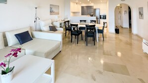 Una sala de estar moderna con un sofá blanco, una mesa de comedor con sillas, una cocina con refrigerador y estufa, y un pasillo que conduce a otra habitación.