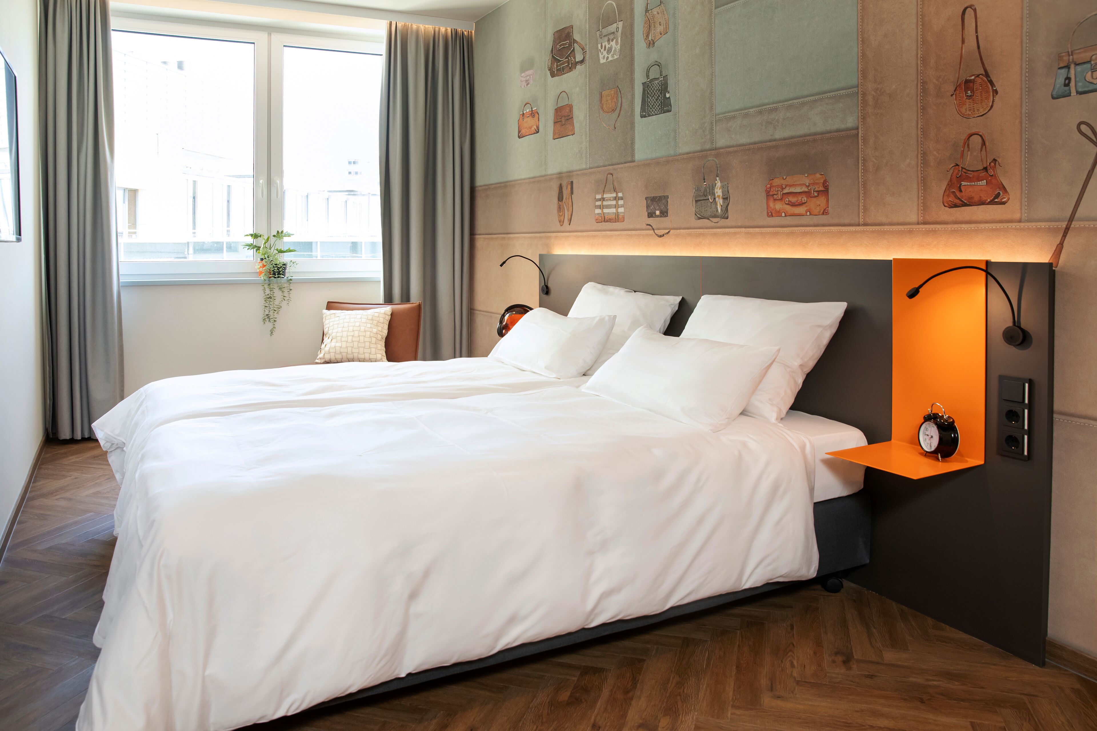 Een tweepersoonsbed met witte lakens, een nachtkastje met een klok en een wandplank met handtassen.