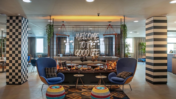 A modern lobby with a checkered column, two blue armchairs, a colorful ottoman, and a neon sign reading "WELCOME TO THE GOOD LIFE".