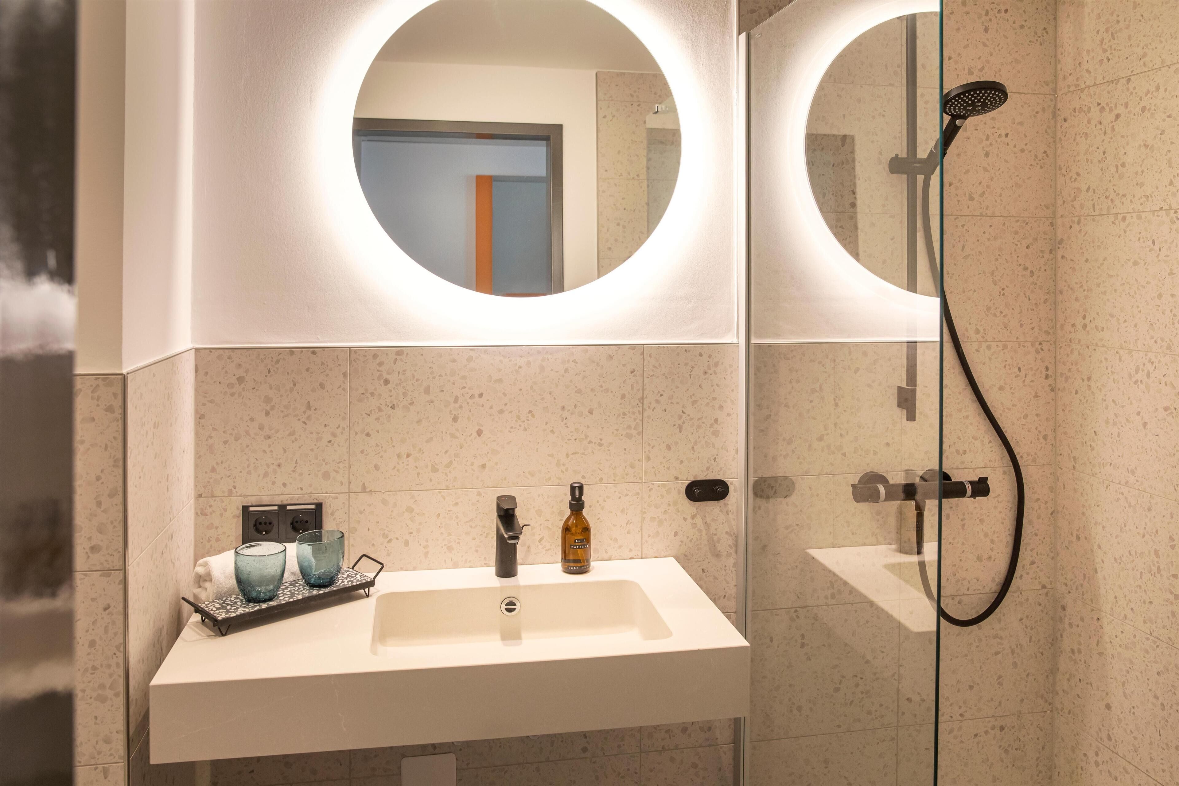 A modern bathroom with a white sink, two round mirrors, a handheld showerhead, and a glass shower enclosure.