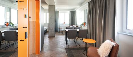 Una habitación de hotel moderna con suelo de madera, una zona de comedor con mesa y sillas, un sofá con un cojín mullido y grandes ventanas con cortinas.