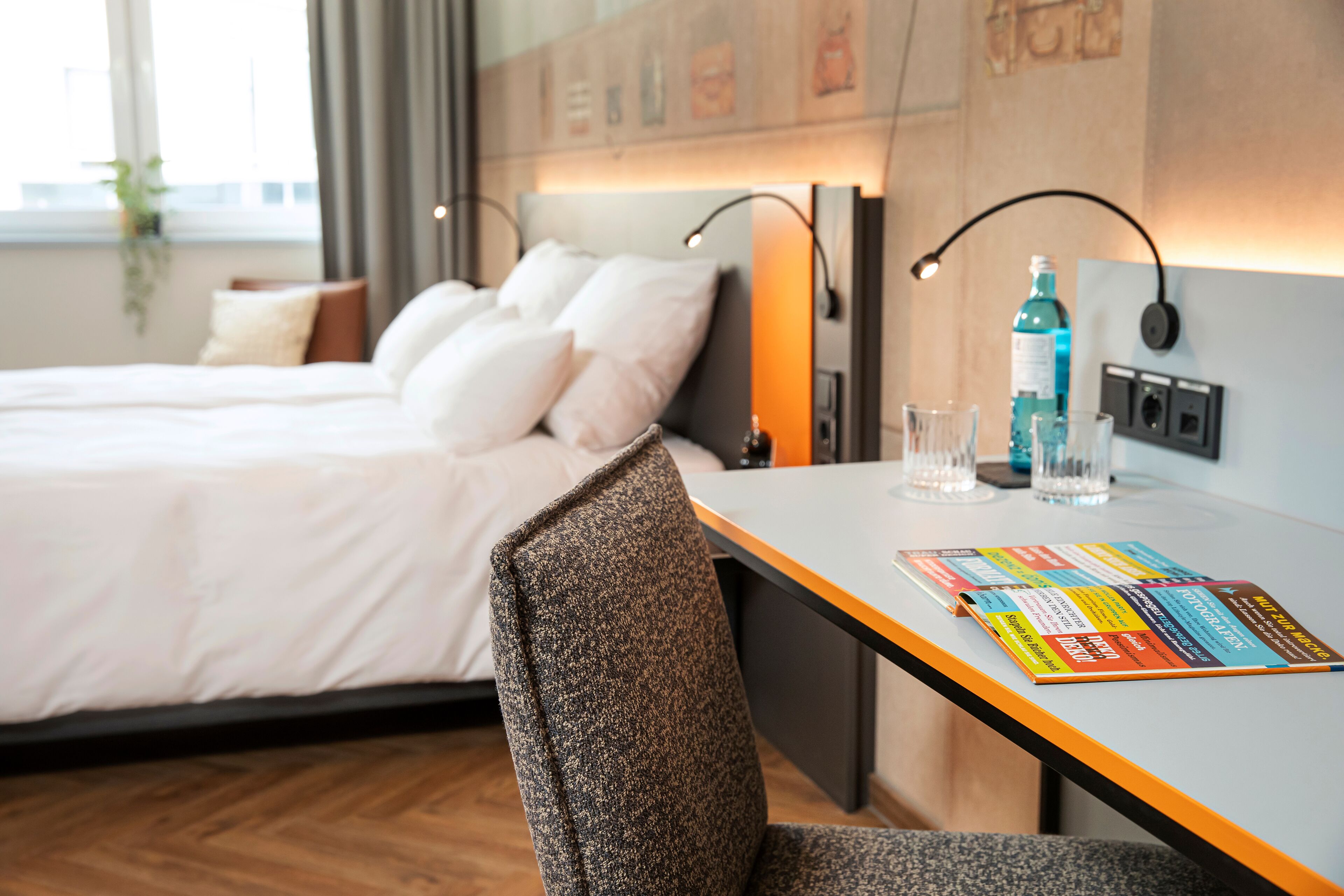 A hotel room with a neatly made bed, a desk with a chair, a wall-mounted lamp, and a bottle of water.