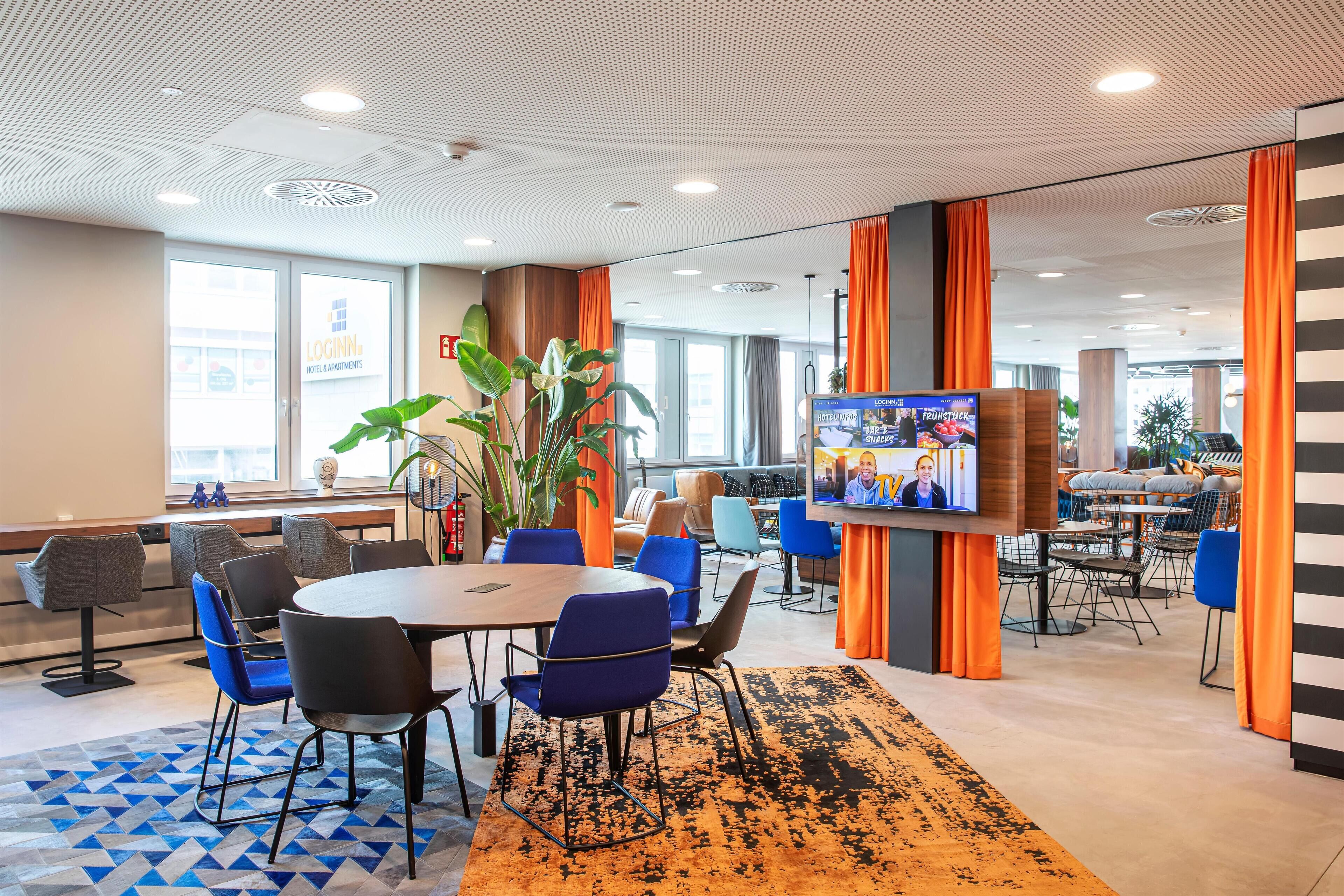 Un moderno vestíbulo de hotel con una mesa redonda, sillas azules, una planta grande y una pantalla de televisión.