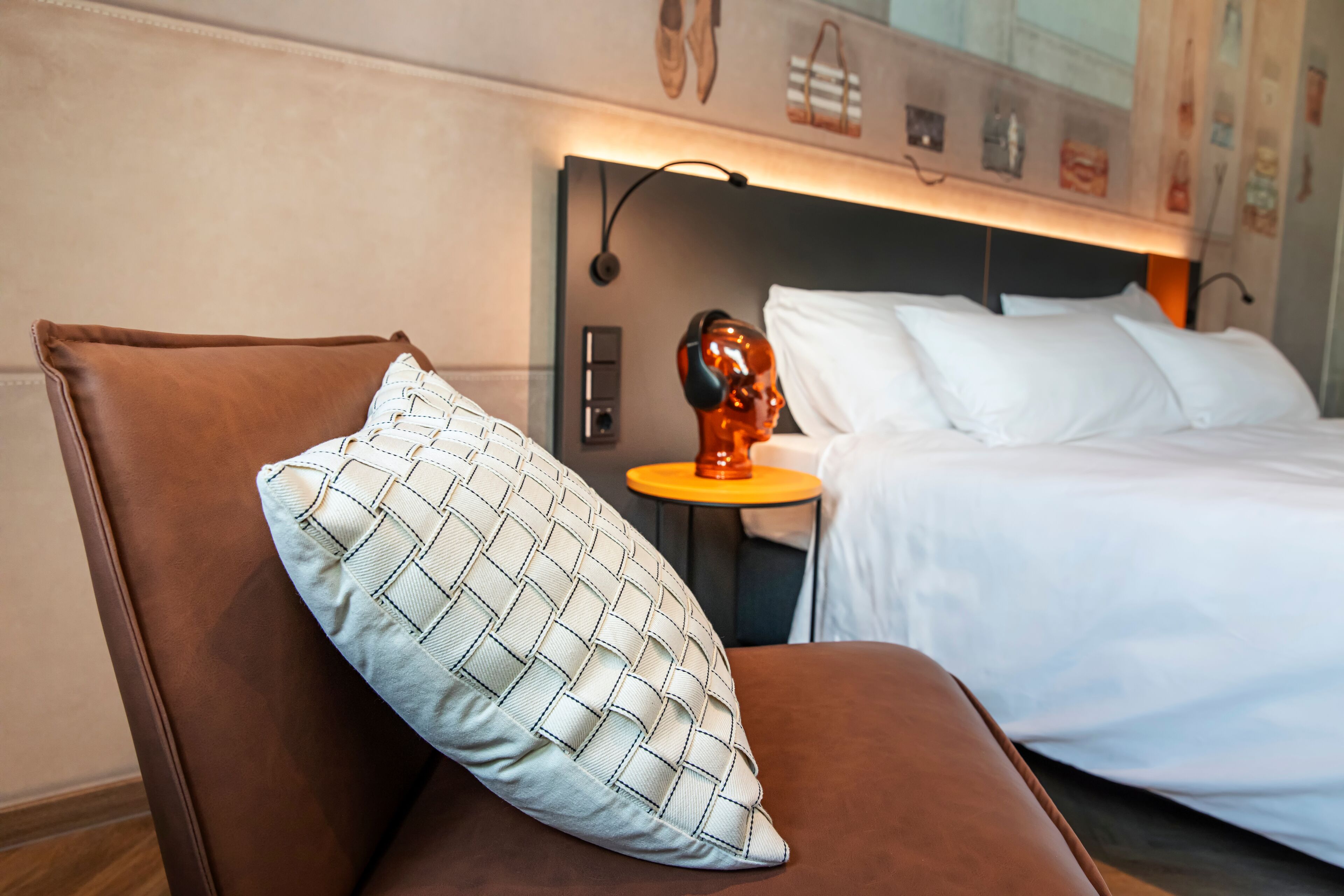 A hotel room with a brown leather chair, a white pillow with a woven pattern, a bed with white linens, a small round table with a red object, and a wall with various pictures and a shoe rack.