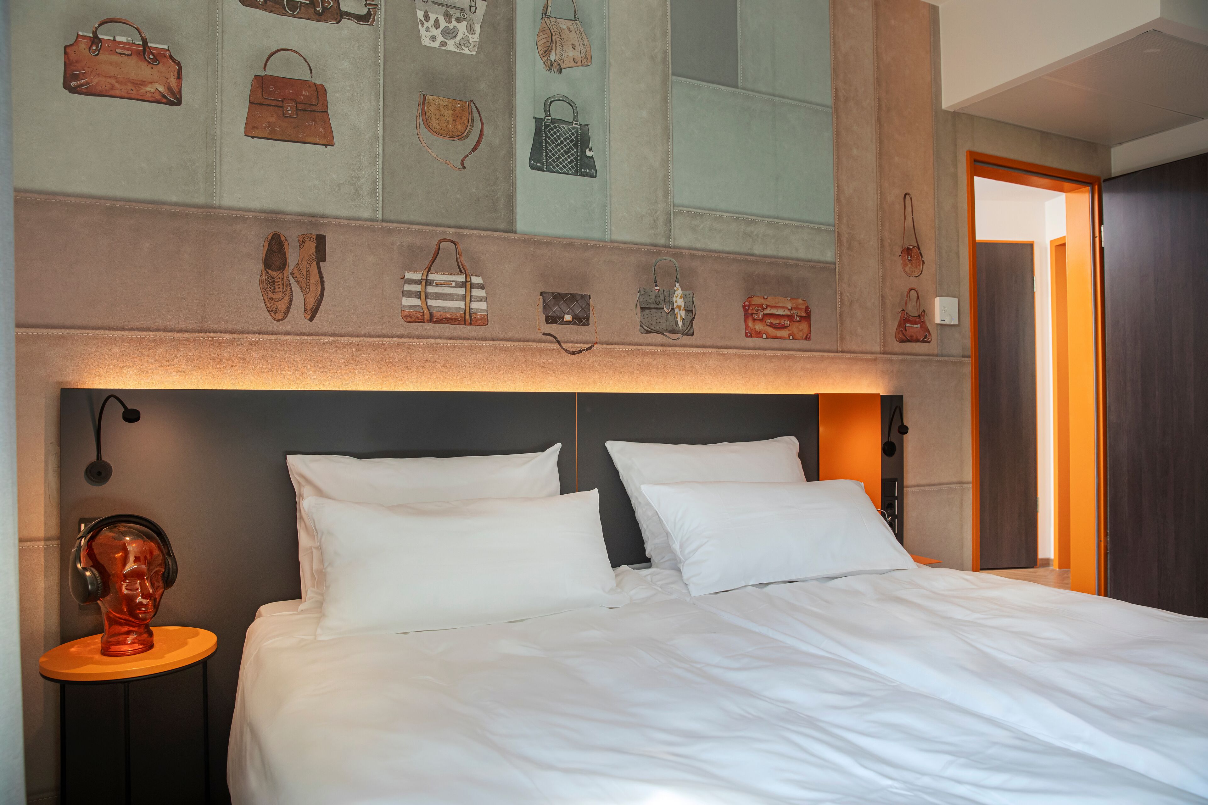 A hotel room with a large bed, white bedding, a headboard with a light fixture, a small table with a sculpture, and a wall decorated with various handbags.