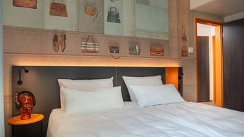 A hotel room with a large bed, white bedding, a headboard with a light fixture, a small table with a sculpture, and a wall decorated with various handbags.