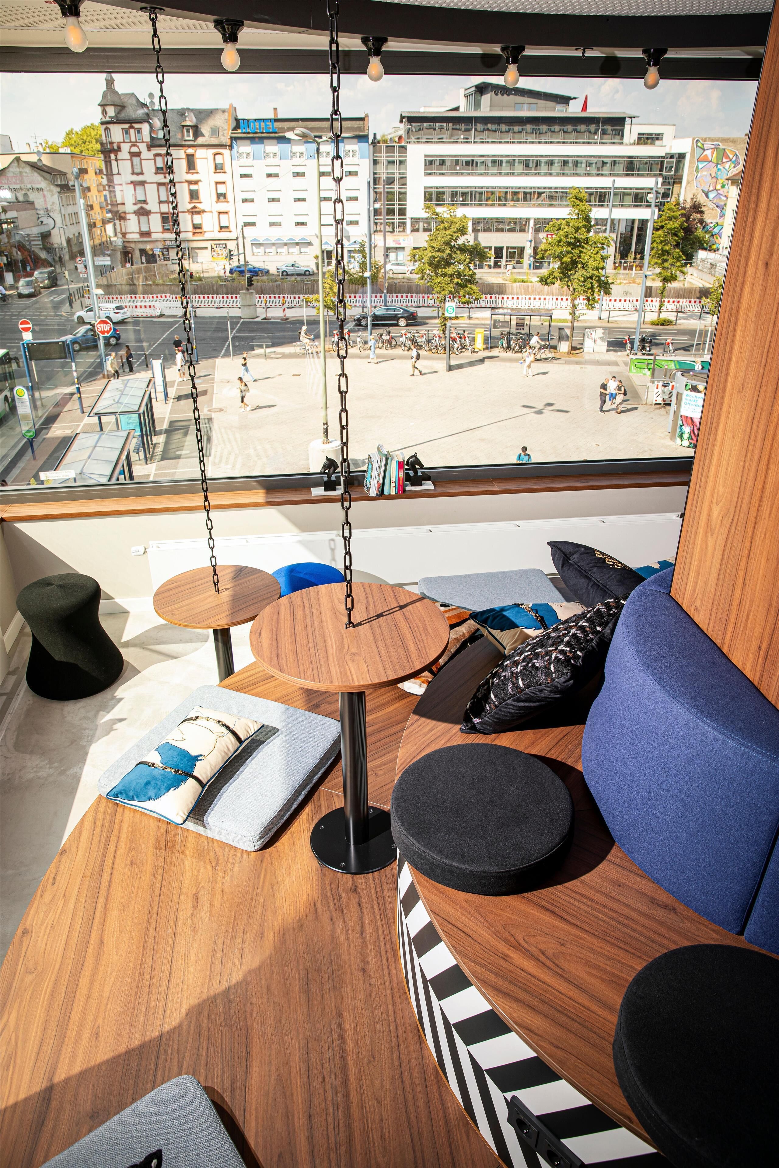 A modern interior with a wooden table, blue seating, and a view of a cityscape through large windows.