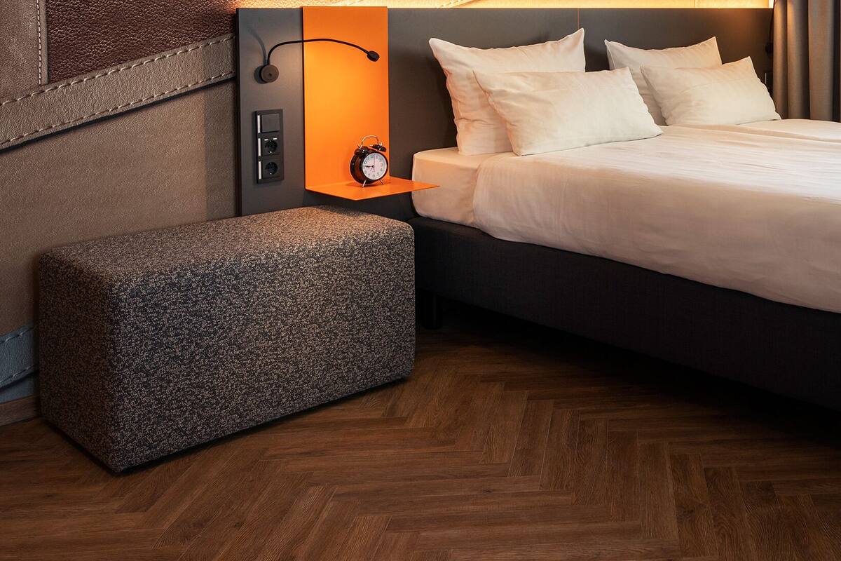 A hotel room with a large bed, a bedside table with a clock, and a small ottoman.