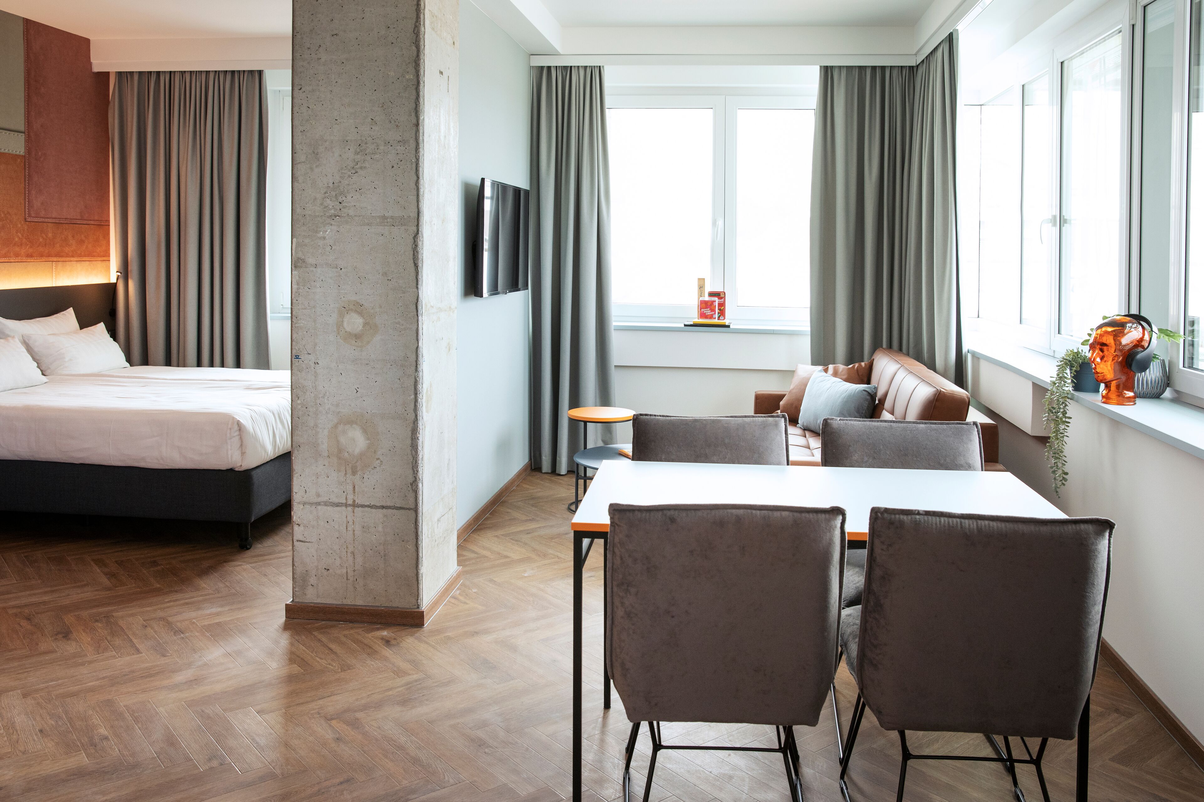 A modern hotel room with a bed, a dining area with a table and chairs, and a large window with curtains.