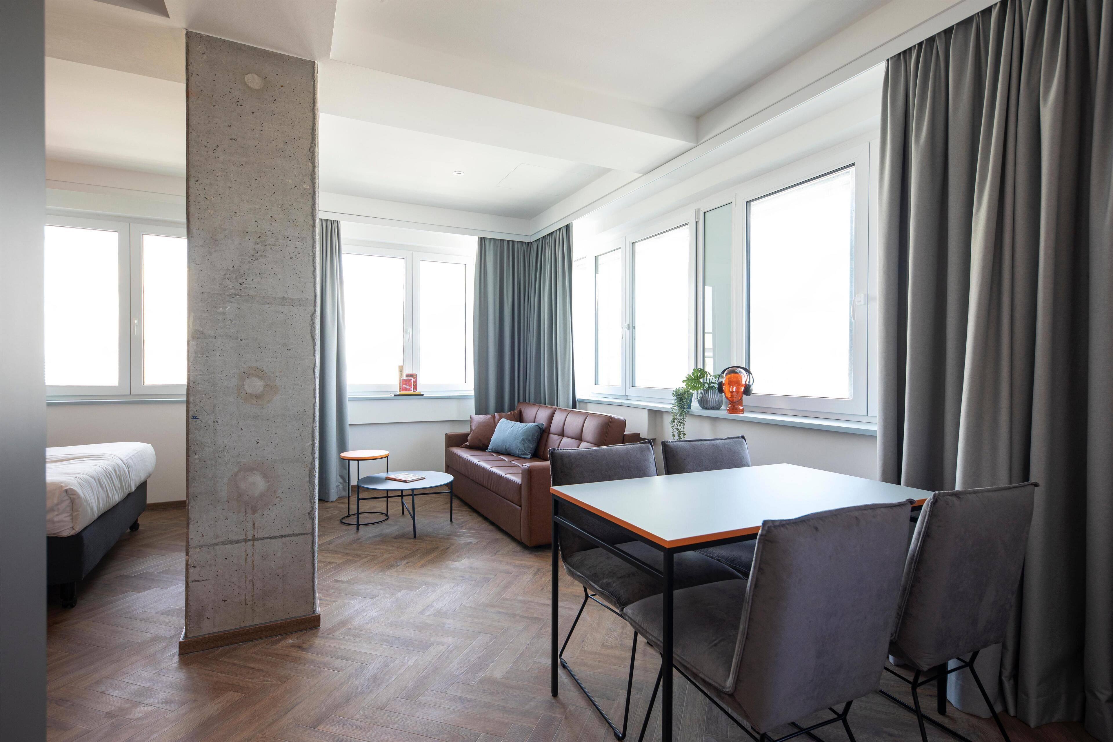 A modern hotel room with a large window, a sofa, a dining table, and a bed.
