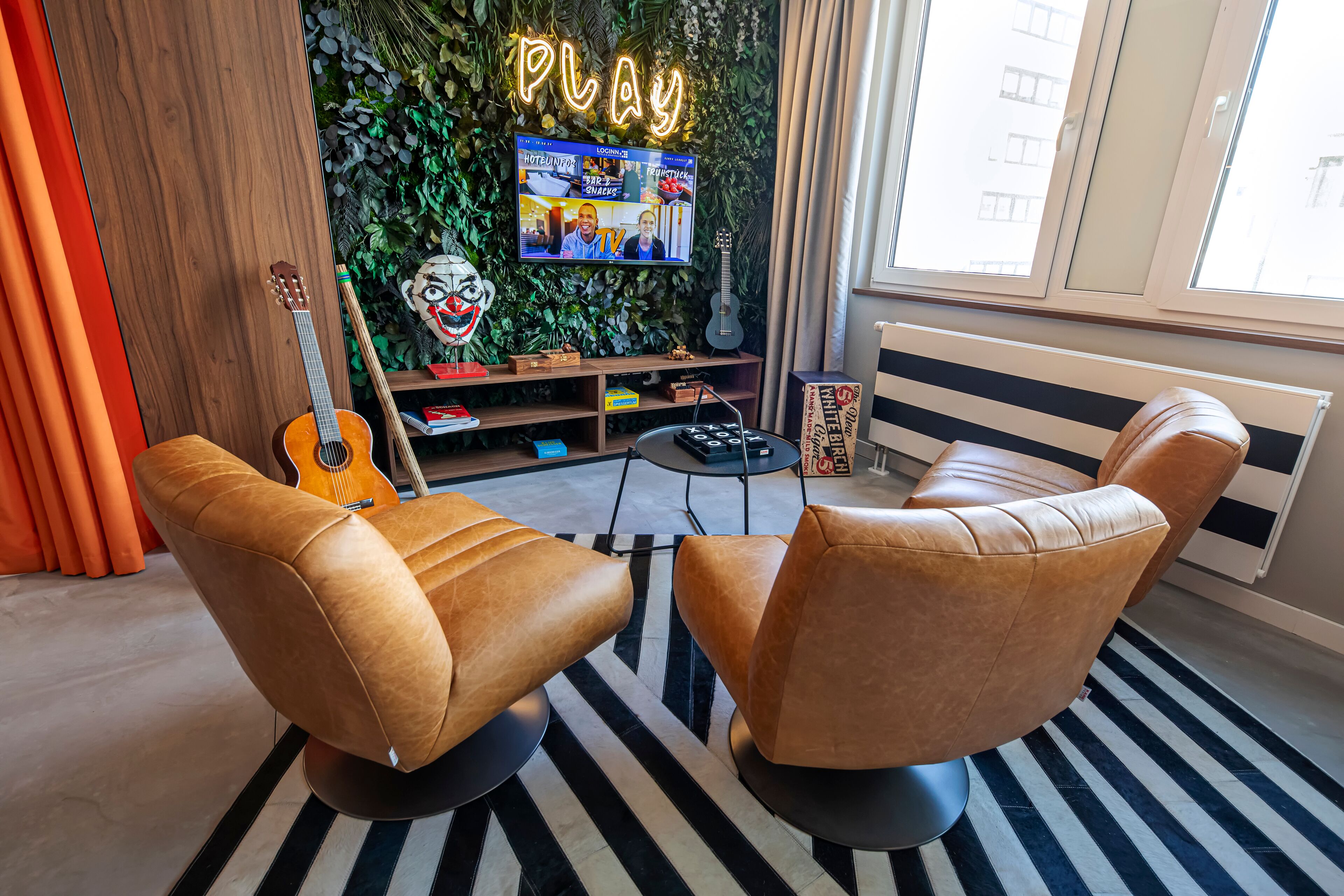 Una sala moderna con dos sillones de cuero, una guitarra, un televisor mostrando un partido y una pared con plantas y un letrero de neón.