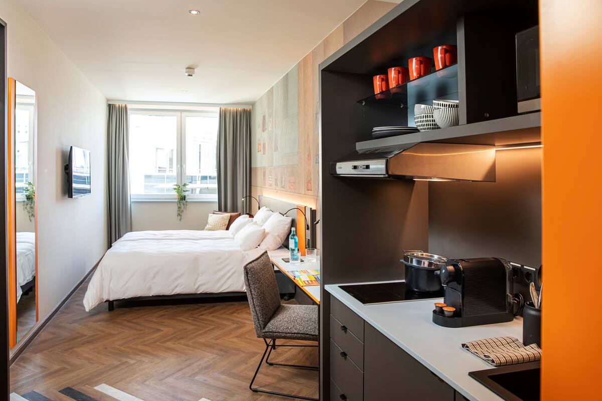 A modern hotel room with a bed, desk, and kitchenette.