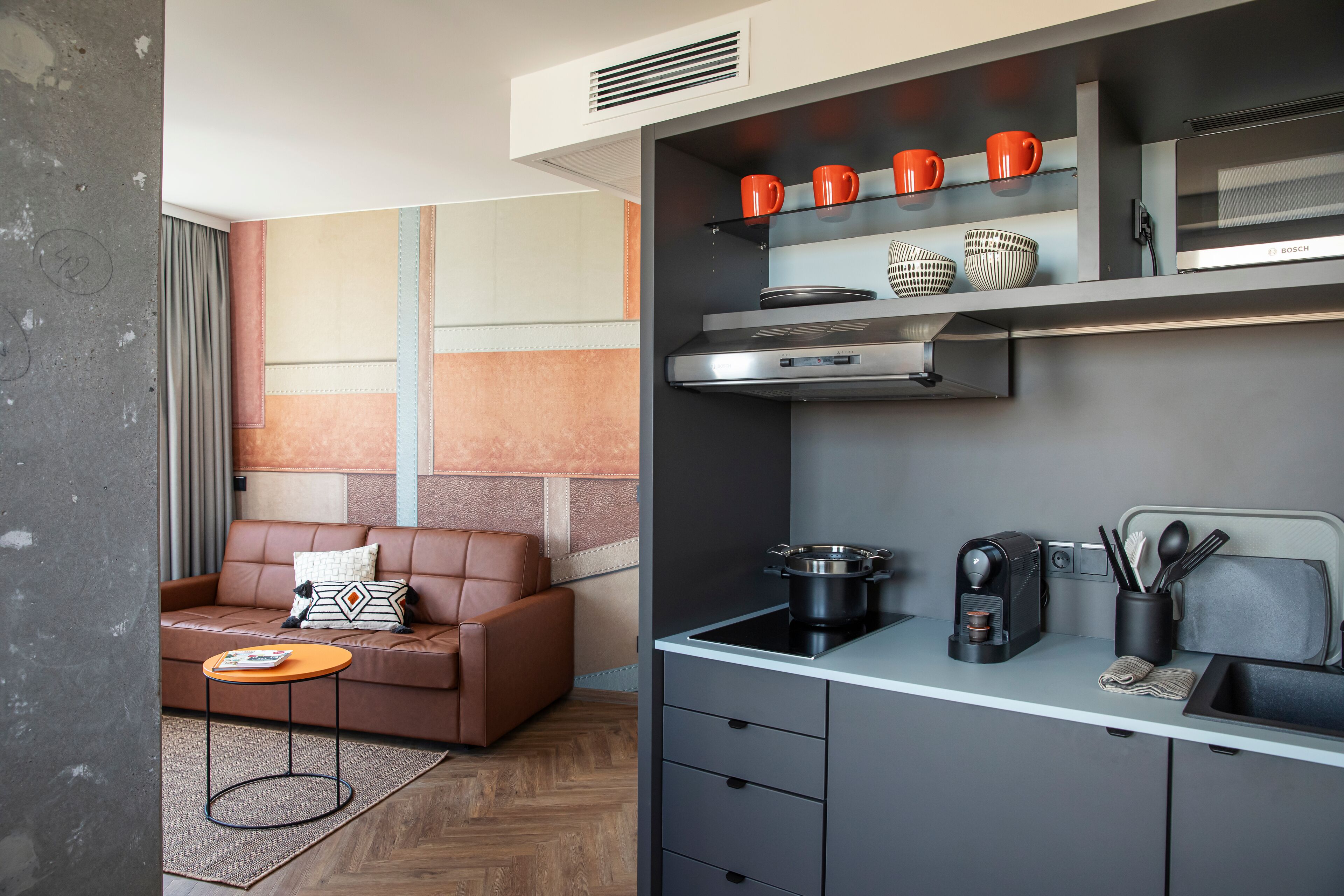 Una cocina moderna con horno Bosch, una cafetera y un lavabo. Hay un sofá de cuero marrón con un cojín estampado en la zona de estar.