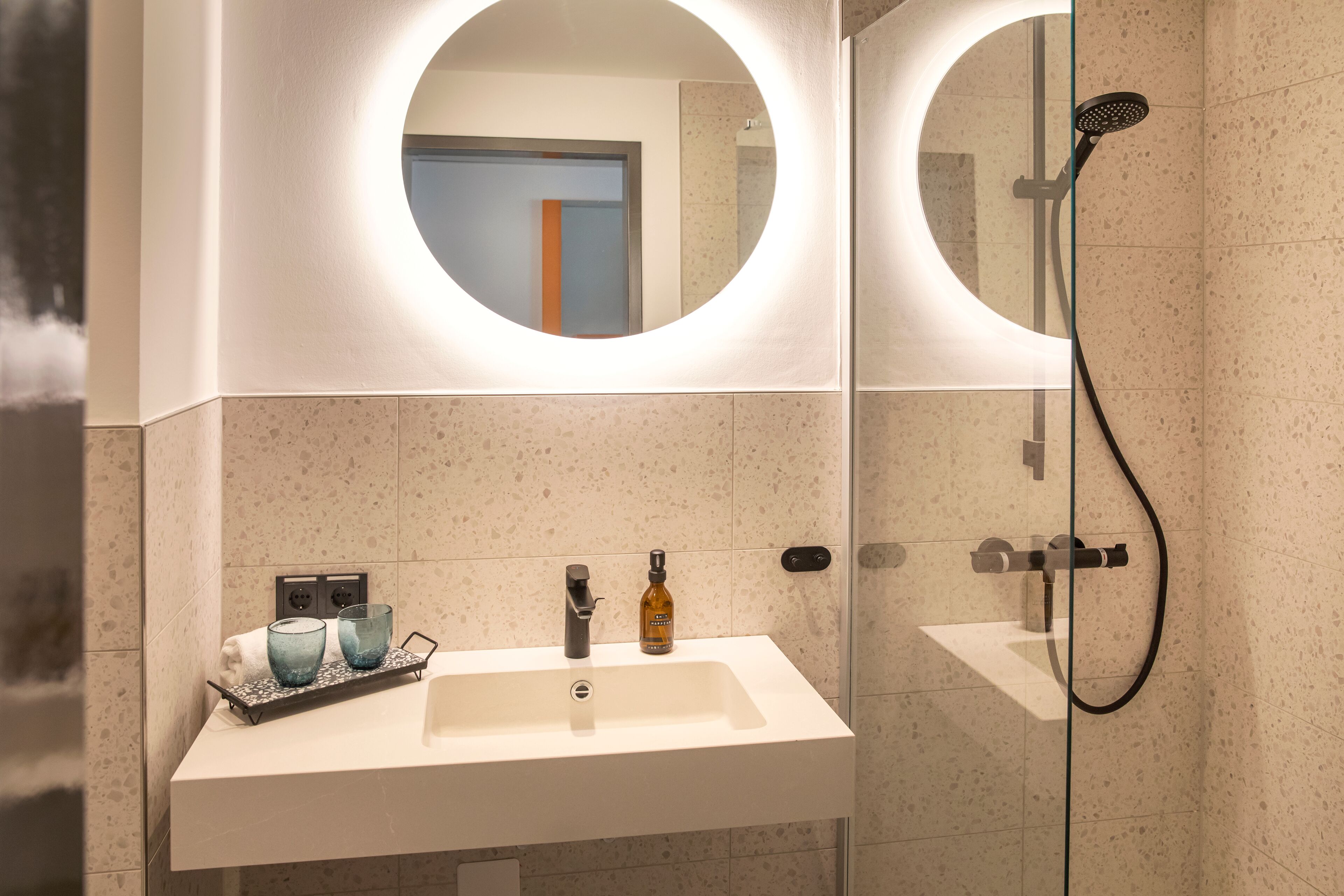 Ein modernes Badezimmer mit einem weißen Waschbecken, zwei runden Spiegeln, einer Duschbrause und einer Glasdusche.