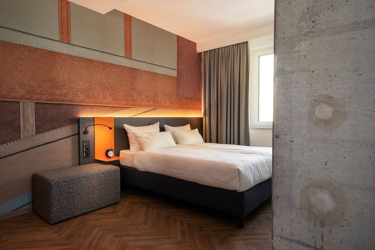 A modern hotel room with a large bed, a bedside table with a lamp, a bench, and a view of the outside.