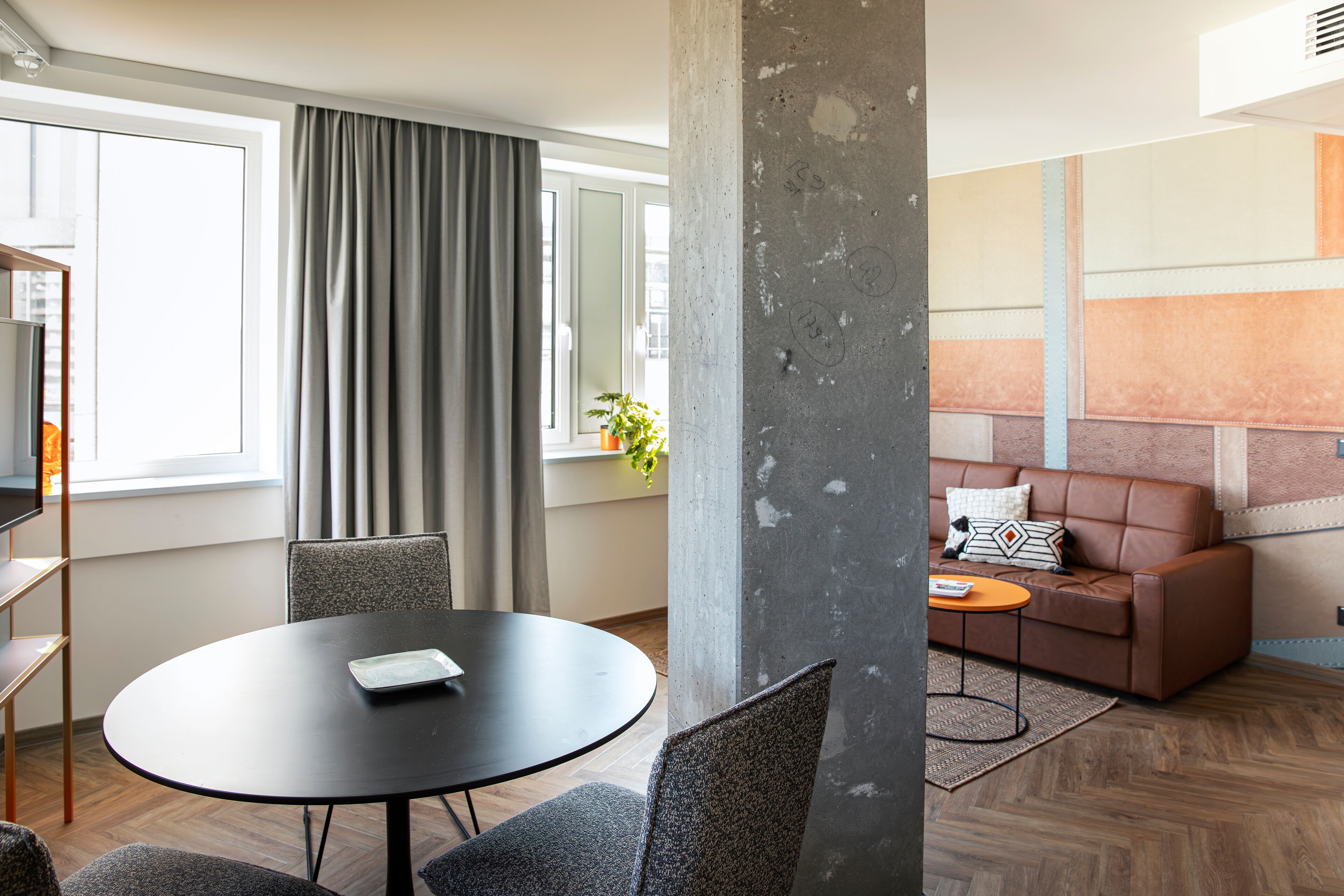 A modern living room with a round table, grey chairs, a brown leather sofa, and a large window with curtains.