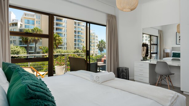 A modern hotel room with a large bed, a desk, and a balcony with a view of high-rise buildings and greenery. - Alexander Road 7B (Cape Town)
