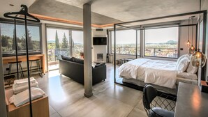 A modern hotel room with a large bed, a sofa, a dining table, and a balcony with a view of the city.
