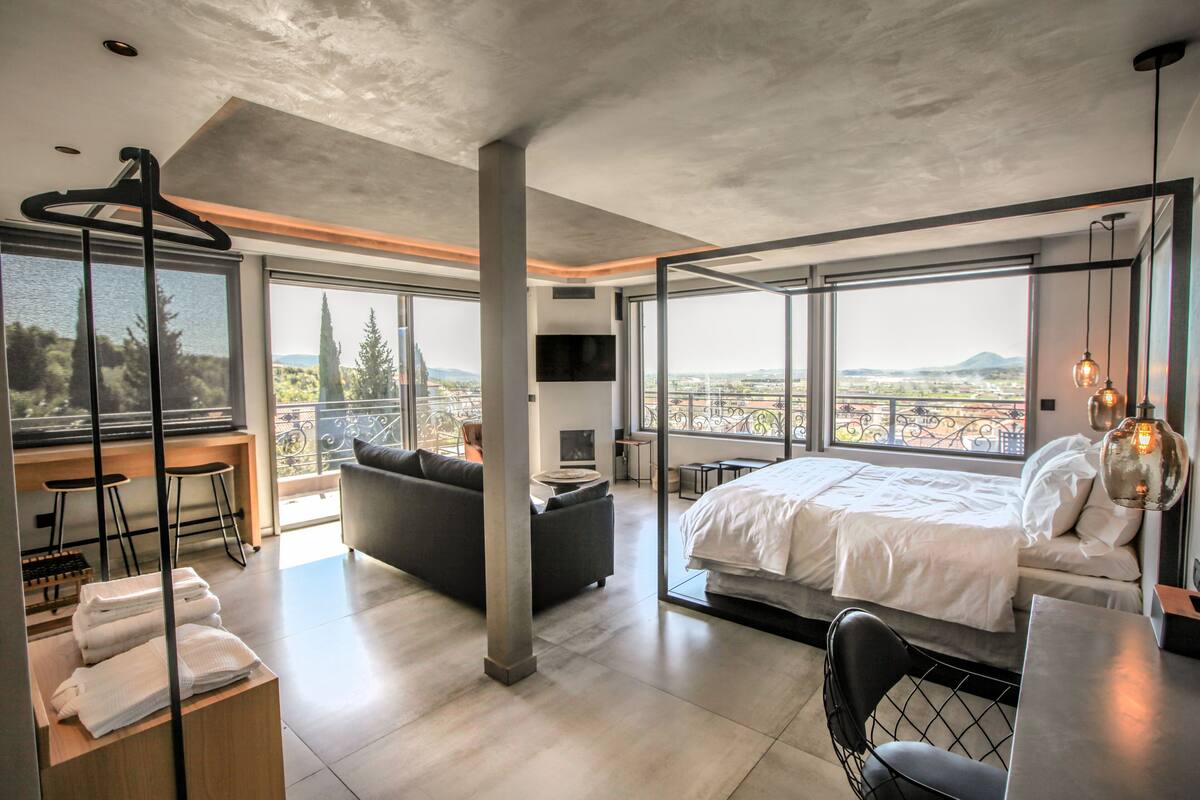 A modern hotel room with a large bed, a sofa, a dining table, and a balcony with a view of the city.