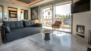 A modern living room with a grey sofa, a round coffee table, and a leather armchair. A fireplace is visible on the right, and a large window offers a view of buildings outside.