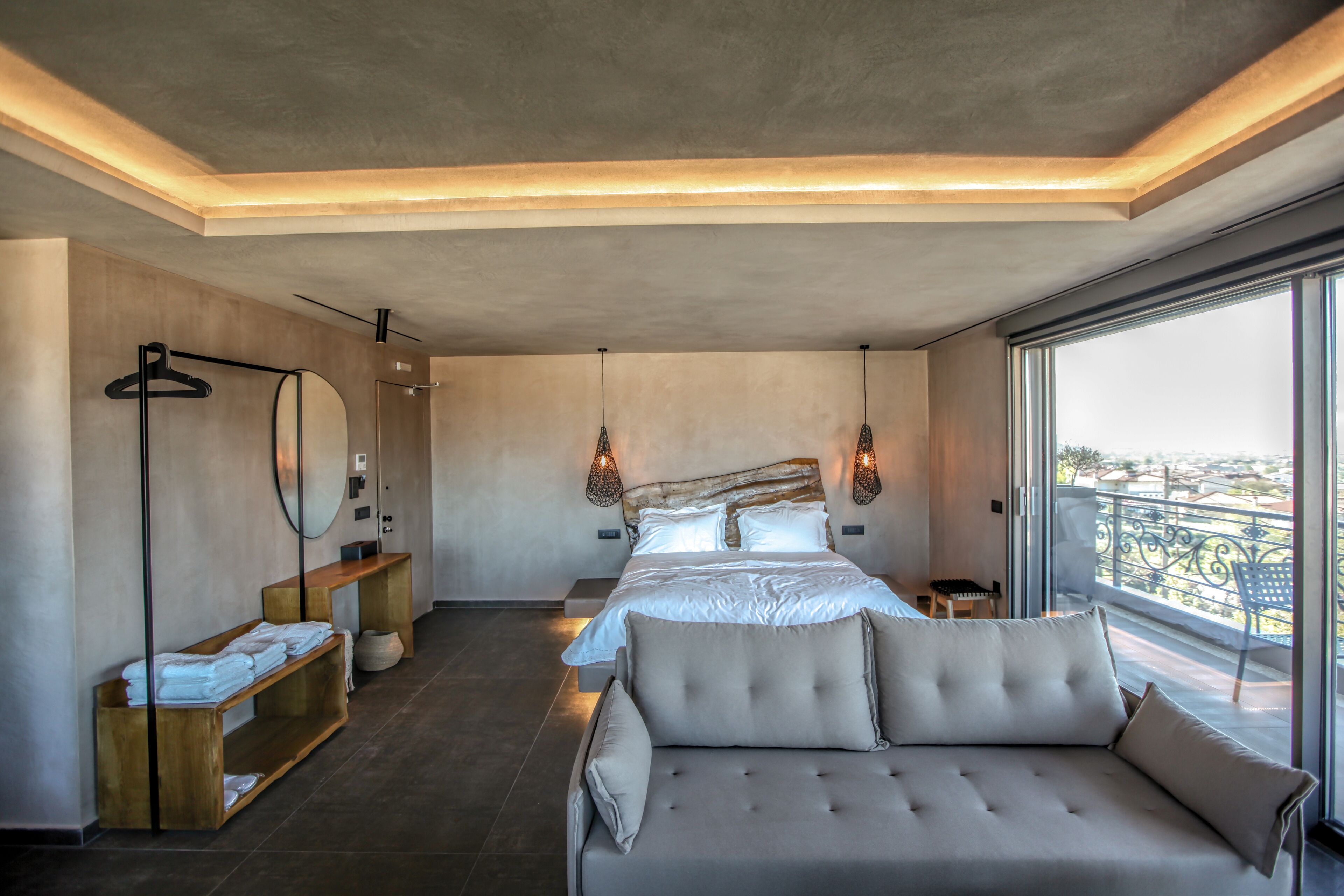 A modern bedroom with a large bed, a sofa, a wooden shelf with towels, a mirror, and a view of the city through a large window.
