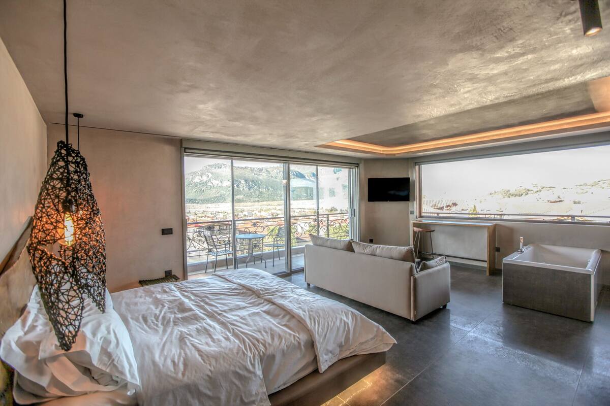 A modern bedroom with a large bed, a sofa, a TV, and a balcony with a view of the mountains.