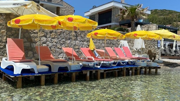 A row of sun loungers with yellow umbrellas, a stone building, and clear water in the foreground. - PALMİYE PANSİYON (Karaburun)