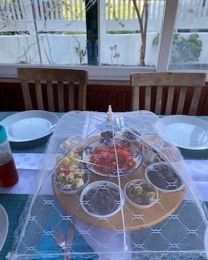 A table set with a variety of dishes covered by a protective mesh, chairs, and a window with a view of the street. - PALMİYE PANSİYON (Karaburun)