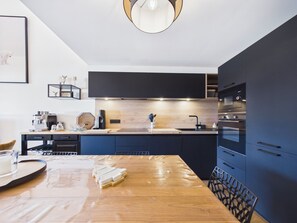A modern kitchen with dark cabinetry, a wooden dining table, and a coffee machine. - Chic Apartment with Balcony, 2 Bedrooms & Mountain Views in Les Gets (Les Gets)