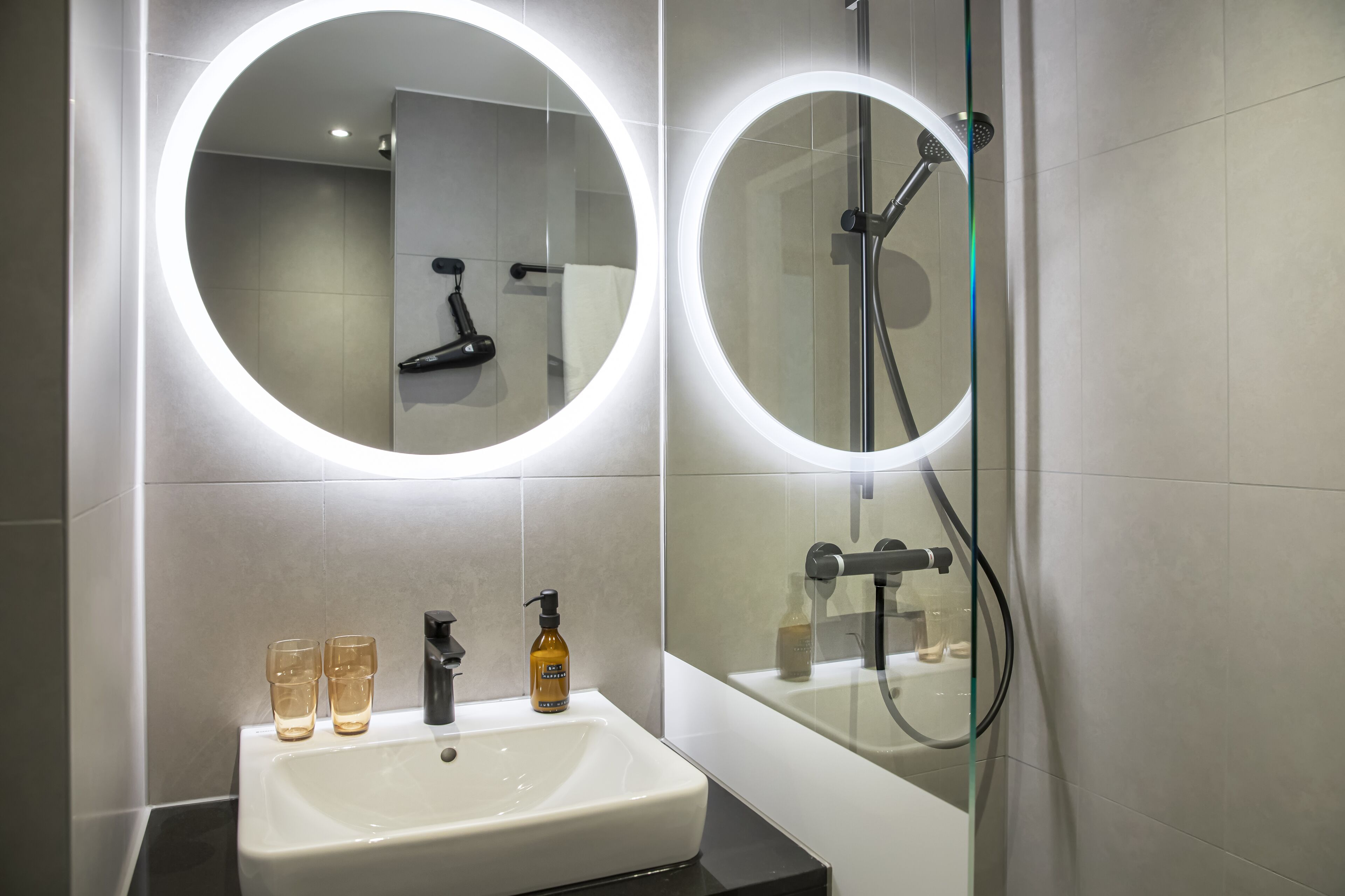 Une salle de bain moderne avec un lavabo blanc, deux verres, un distributeur de savon et un miroir doté d’un éclairage circulaire.