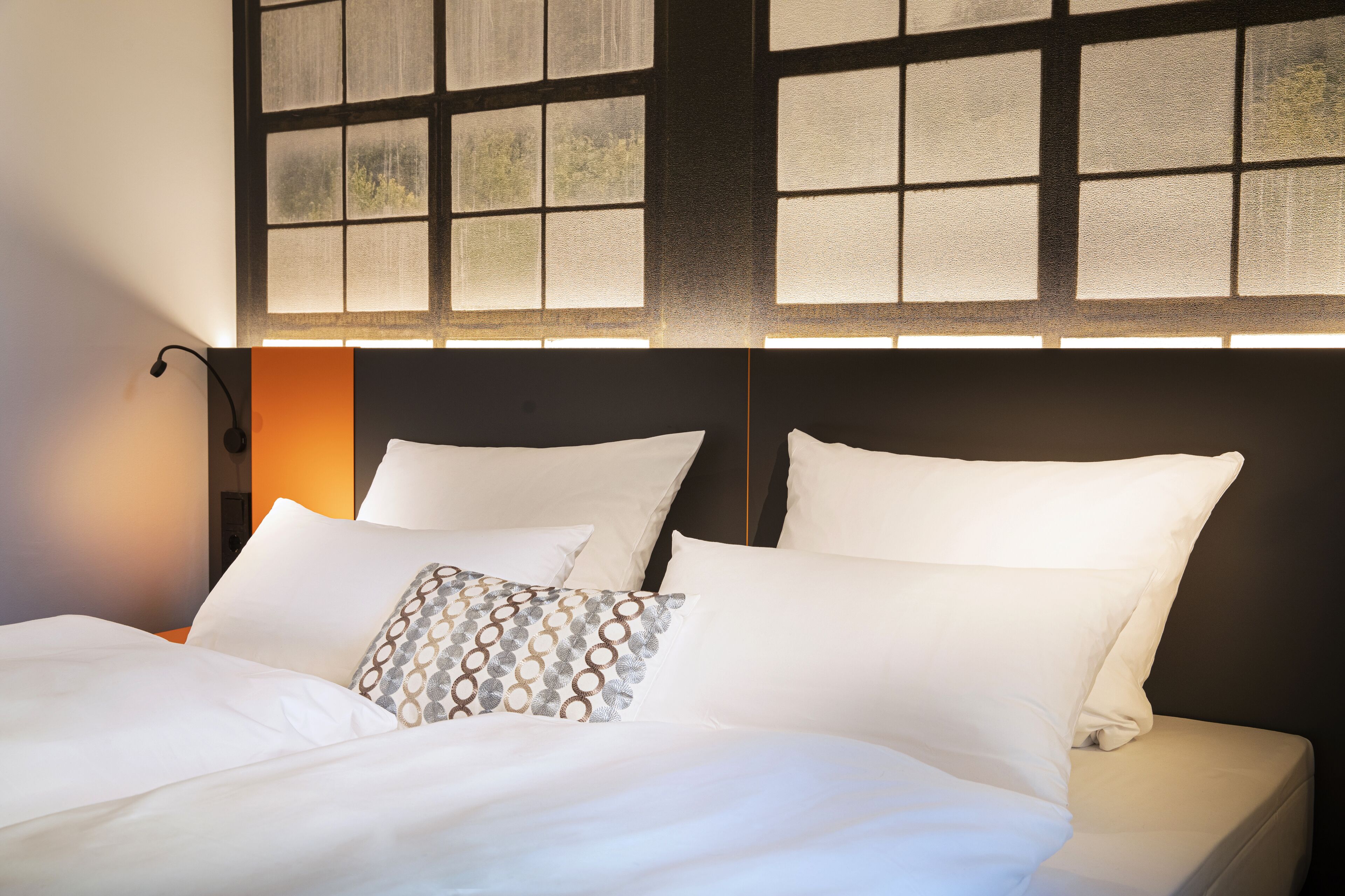 Una cama bien hecha con sábanas blancas y almohadas estampadas, una lámpara de noche y vistas a través de una ventana con diseño de cuadrícula.