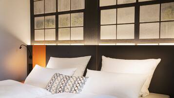 Una cama bien hecha con sábanas blancas y almohadas estampadas, una lámpara de noche y vistas a través de una ventana con diseño de cuadrícula.