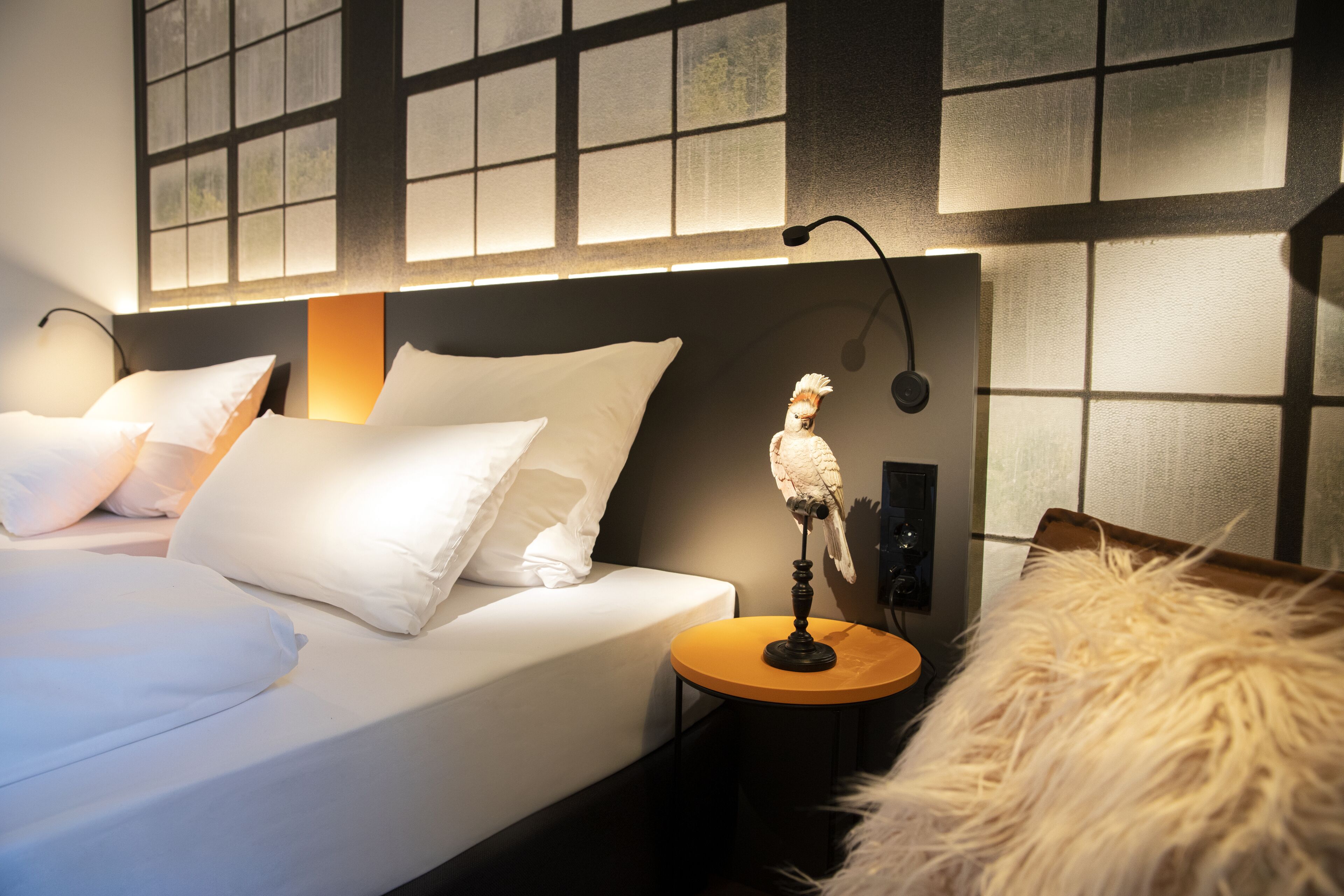 A neatly made bed with white linens and pillows, a small round table with a lamp, and a decorative bird statue.