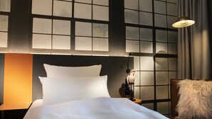 A hotel room with a large bed, a nightstand, a lamp, and a view through a window with a grid pattern. - Loginn Hotel Waiblingen (Waiblingen)