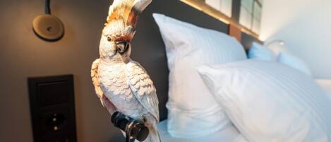 Un oiseau était perché sur un support dans une pièce où se trouvaient un lit et un luminaire fixé au mur.