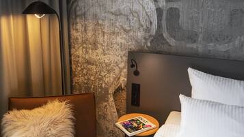 Habitación de hotel con un gran mural en la pared de un cacatúa, un sillón de cuero marrón con un cojín mullido, una mesita de noche con una revista y una cama bien hecha con ropa de cama blanca.