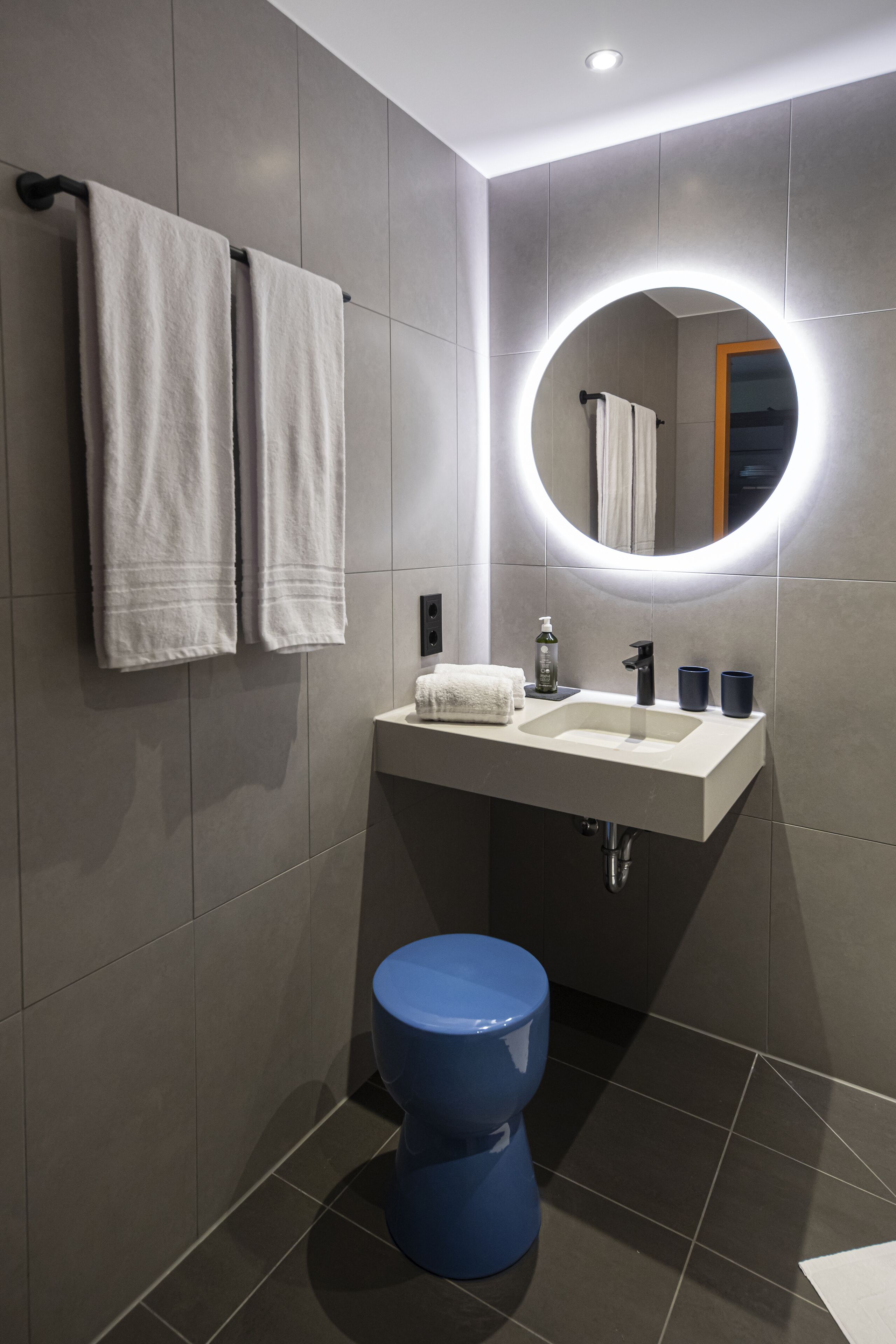 Ein modernes Badezimmer mit einem weißen Waschbecken, einem runden Spiegel und einer blauen Toilette.