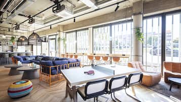A modern lounge area with a large table, comfortable seating, and large windows offering a view of the street.
