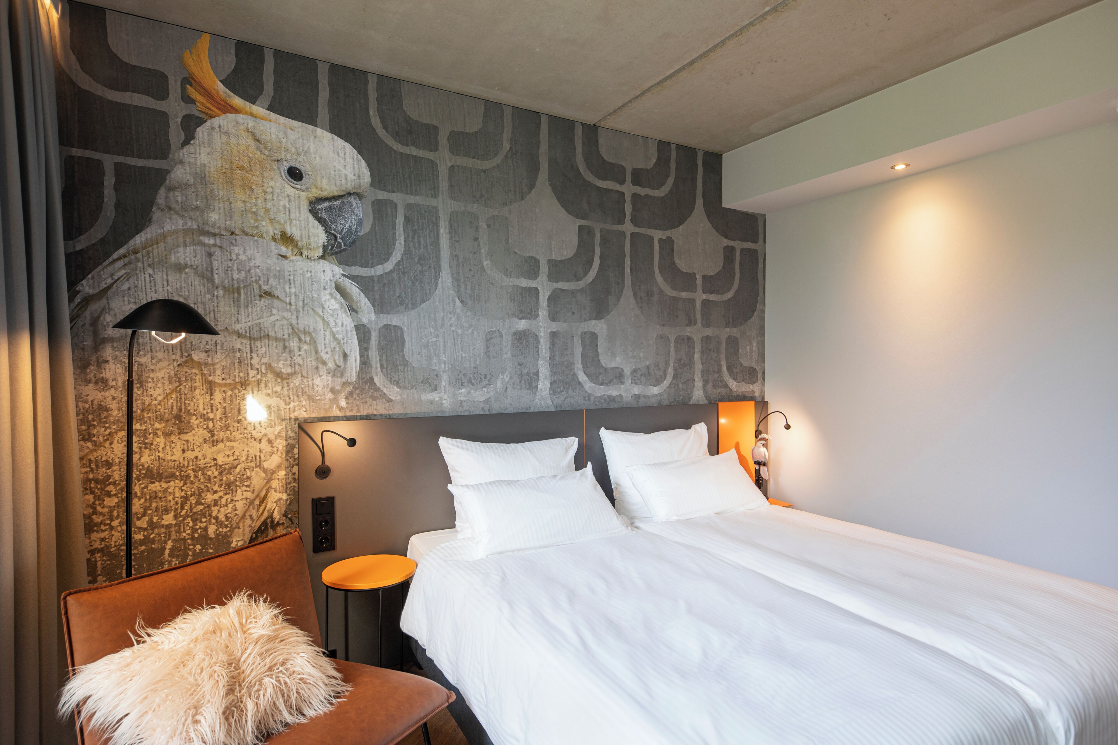 Habitación de hotel con un gran mural en la pared de un cacatúa, una cama con ropa blanca, una silla marrón con un cojín mullido y una mesita redonda pequeña.