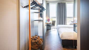 A hotel room with a bed, a desk with a chair, a window with curtains, a coat rack, and a backpack on a bench.