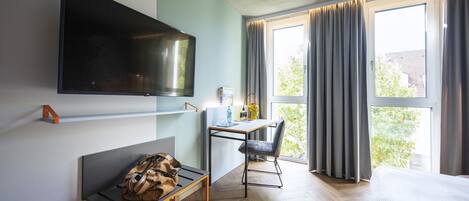 A hotel room with a bed, a desk with a chair, a TV mounted on the wall, and a window with curtains.