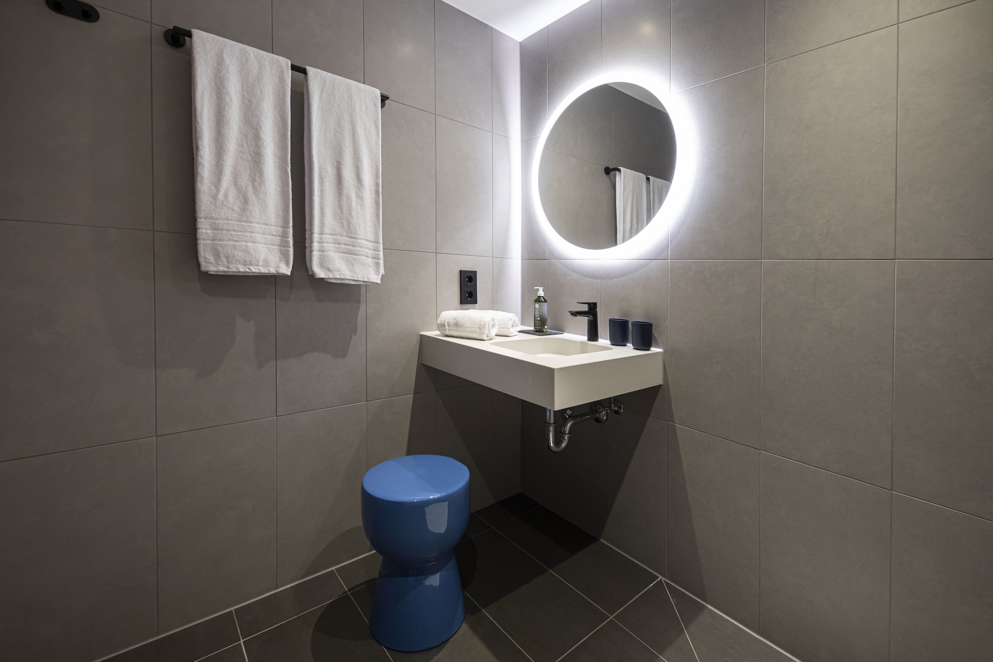 Een moderne badkamer met grijze tegels, een ronde spiegel met verlichting, een wit wastafel, een blauw toilet en twee witte handdoeken aan de muur.