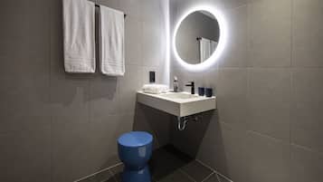 A modern bathroom with grey tiles, a round mirror with a light, a white sink, a blue toilet, and two white towels hanging on the wall.
