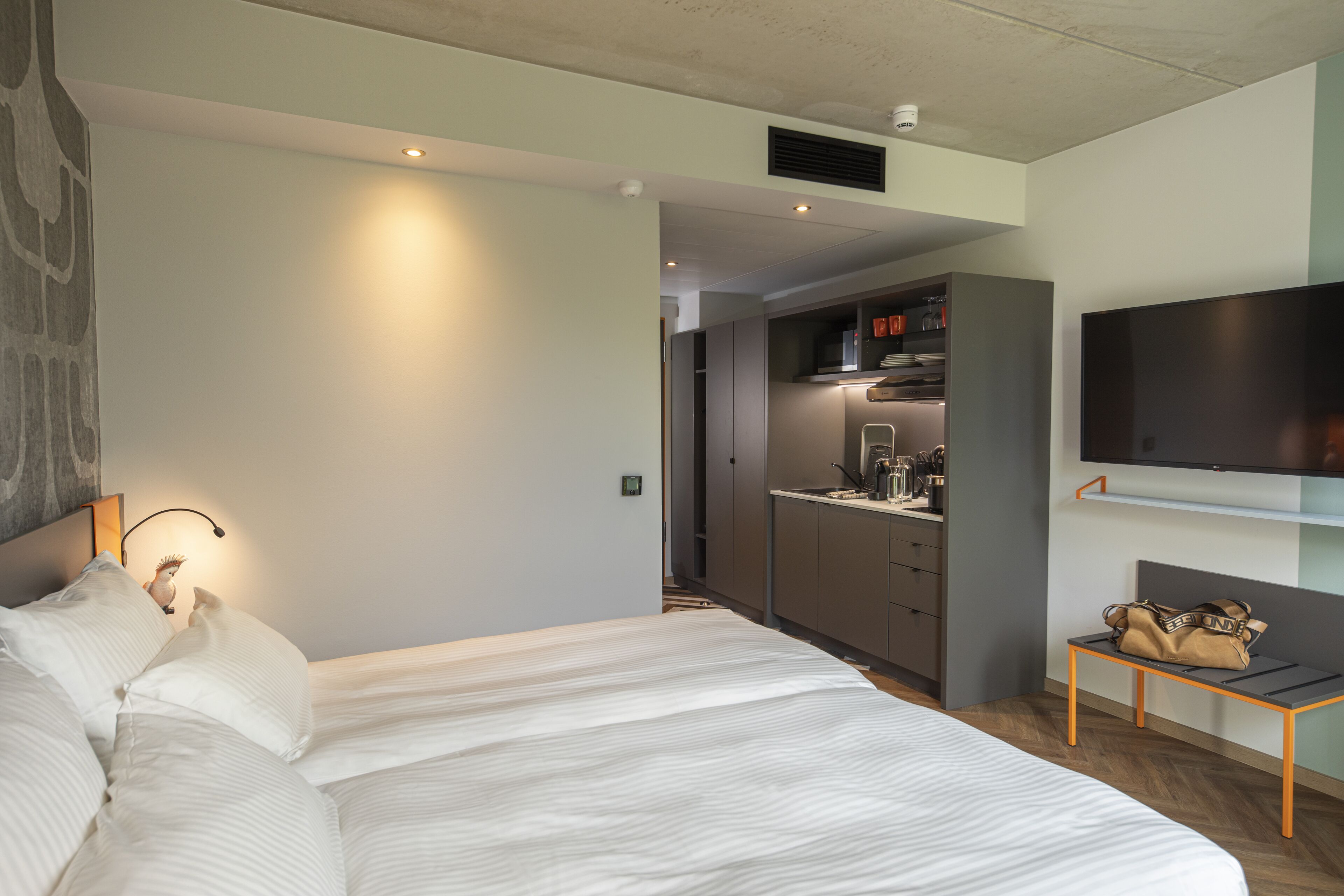 Una habitación de hotel moderna con una cama grande, una mesita con un bolso, un televisor montado en la pared y una zona de kitchenette.