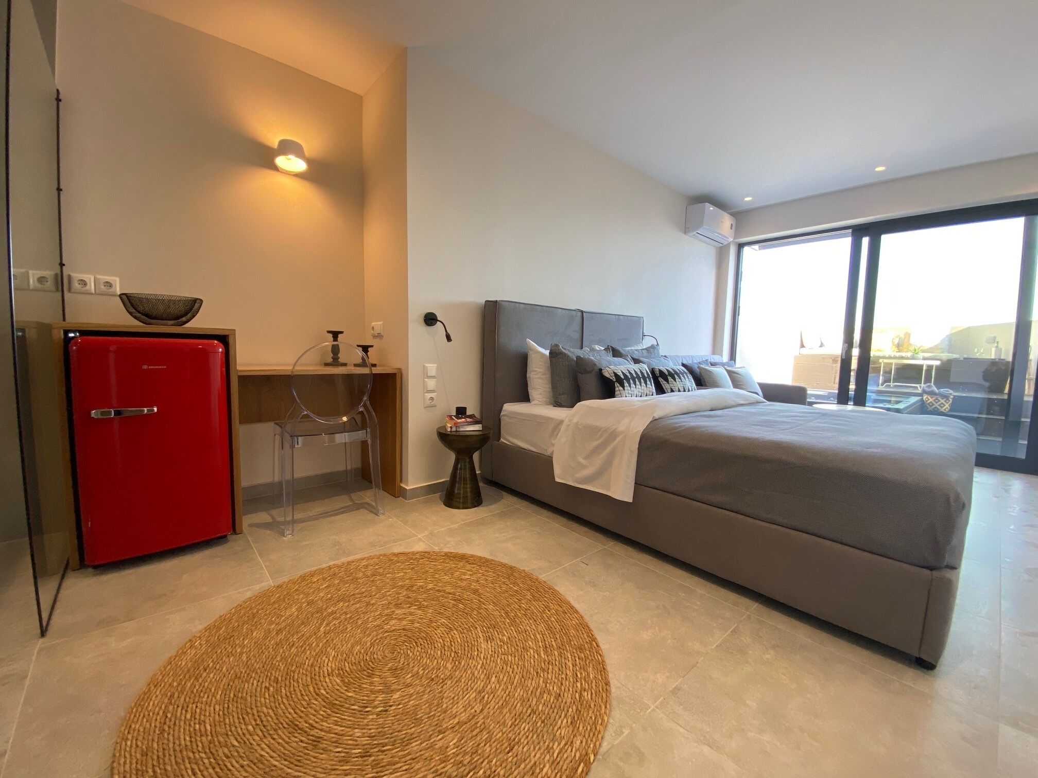 A modern bedroom with a large bed, a red refrigerator, a small table with a chair, and a large window offering a view of the outdoors.